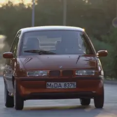Pomarańczowy BMW E1 jedzie po drodze, światła odbijają się od karoserii.