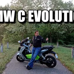Mężczyzna siedzi na skuterze BMW C Evolution, podłączonym do ładowania. Tło to malowniczy krajobraz z drzewami.