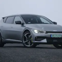 Szary Kia EV6 GT na mglistej drodze, z zielonymi polami i drzewami w tle.