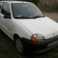 Białe seicento elettra z niebieskim wnętrzem, zaparkowane na tle drewnianego płotu i trawy.