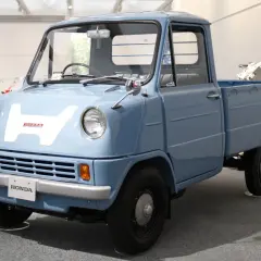 Niebieski, zabytkowy pick-up Honda z dużym białym "H" na masce. Ten model, choć nie jest Isuzu, przypomina klasyczne japońskie pojazdy użytkowe.