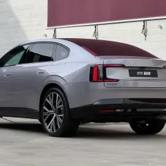 Elegancki, szary Volvo ES90 z charakterystycznymi światłami LED stoi na tle białej, drewnianej ściany.