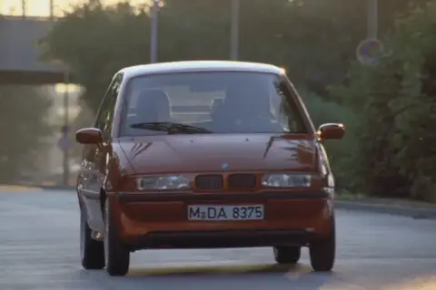 Pomarańczowy BMW E1 jedzie po drodze, światła odbijają się od karoserii.