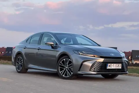 Szary Toyota Camry 2.5 Hybrid na tle zachodzącego słońca i domów.