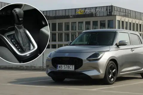 Srebrny Suzuki Swift Hybrid na parkingu przed budynkiem Poczty Polskiej. Widoczny detal dźwigni zmiany biegów.