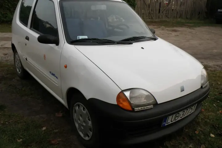 Białe seicento elettra z niebieskim wnętrzem, zaparkowane na tle drewnianego płotu i trawy.