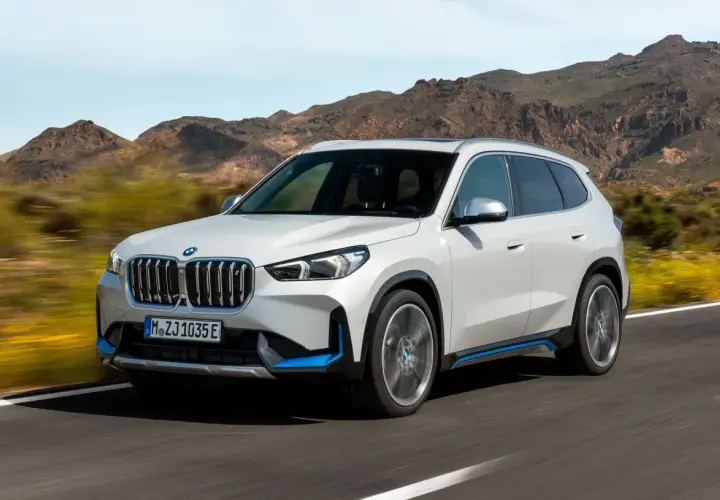 Biały ix1 BMW z niebieskimi akcentami jedzie po drodze, w tle góry.