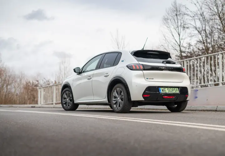 Biały Peugeot e-208 z zieloną tablicą rejestracyjną WE 5G959 zaparkowany na drodze.