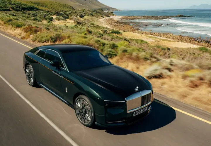 Elegancki, ciemnozielony Rolls-Royce elektryczny sunie po malowniczej drodze nad oceanem.
