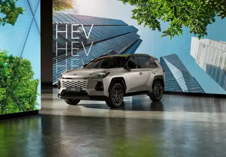 Srebrny SUV RAV4 HEV stoi przed ekranem z nowoczesnymi budynkami i zielenią.