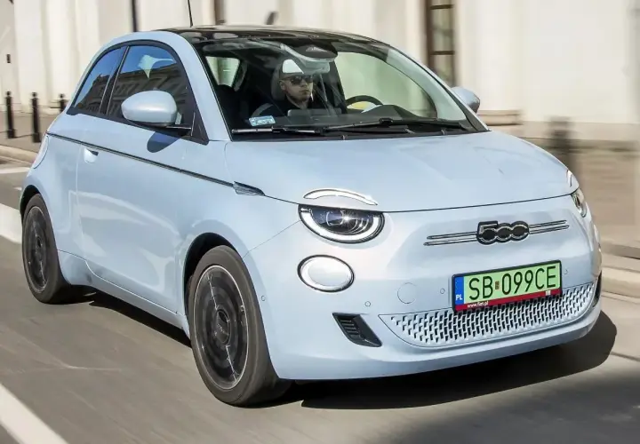 Jasnoniebieski fiat elektryczny 500 z czarnym dachem jedzie ulicą. Kierowca w okularach przeciwsłonecznych prowadzi auto.