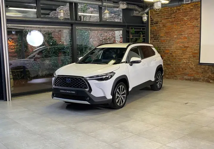 Biała Toyota Corolla Cross Hybrid stoi w nowoczesnym wnętrzu z ceglaną ścianą i dużymi oknami.