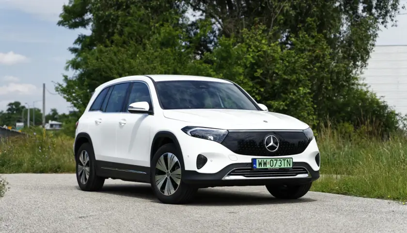 Biały Mercedes EQB na tle zieleni i nieba. Elektryczny SUV z charakterystycznym grillem i nowoczesnymi światłami.