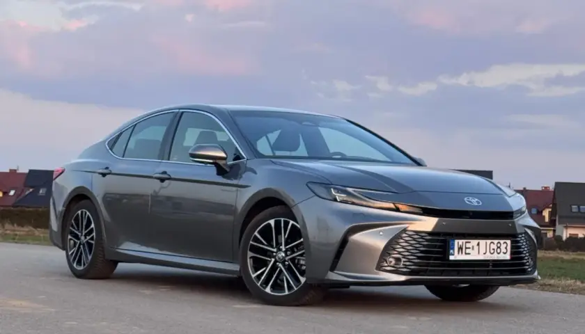 Szary Toyota Camry 2.5 Hybrid na tle zachodzącego słońca i domów.