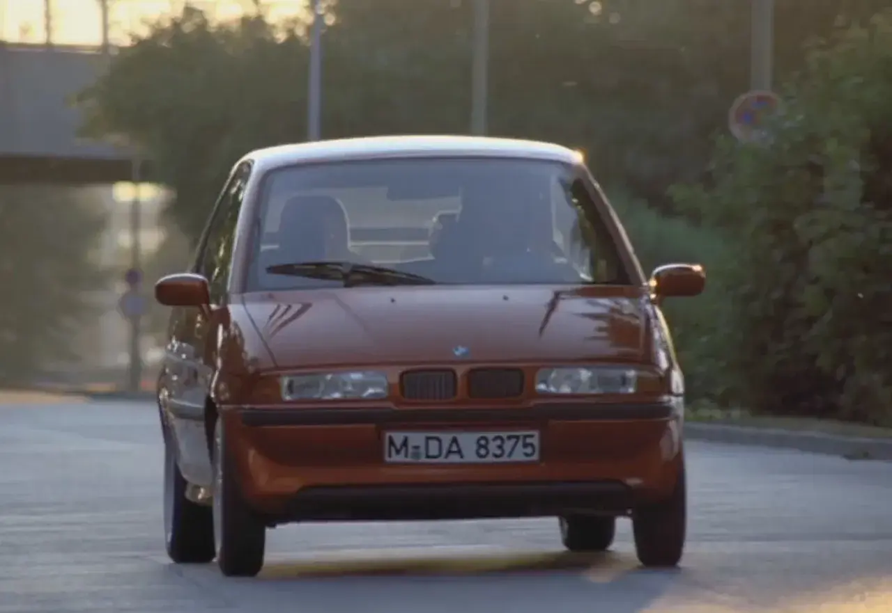 Pomarańczowy BMW E1 jedzie po drodze, światła odbijają się od karoserii.