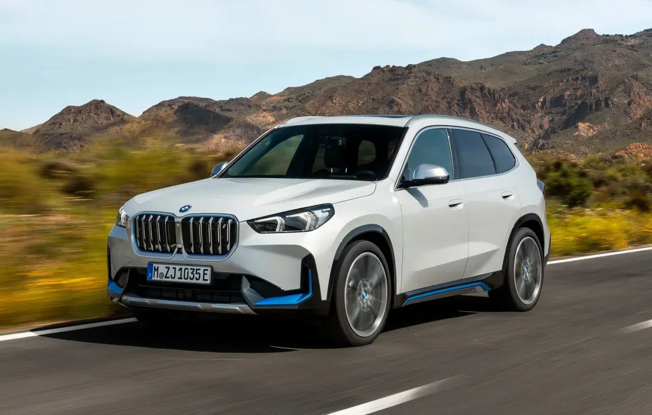 Biały ix1 BMW z niebieskimi akcentami jedzie po drodze, w tle góry.