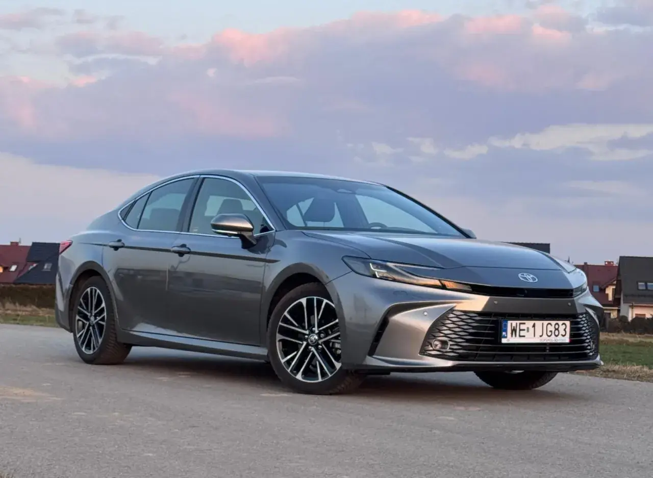 Szary Toyota Camry 2.5 Hybrid na tle zachodzącego słońca i domów.