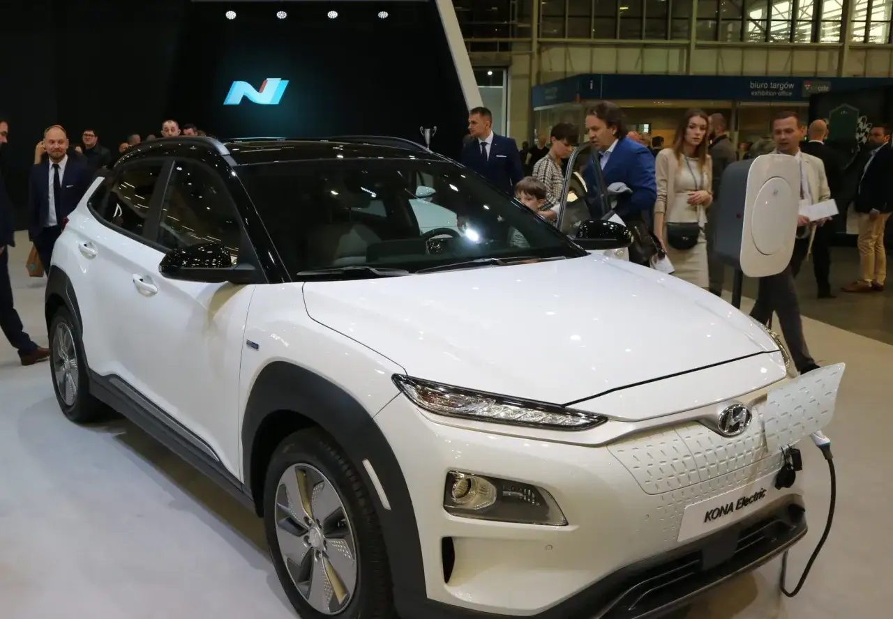 Biały Hyundai Kona Electric na wystawie, podłączony do ładowarki. Ludzie oglądają nowy model.