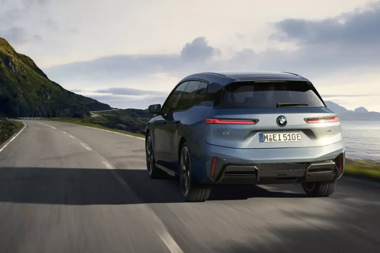 Niebieskie BMW iX jedzie krętą drogą wzdłuż malowniczego wybrzeża.