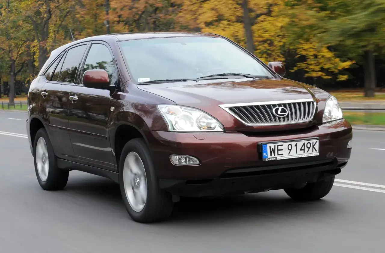 Brązowy Lexus RX 400h jedzie po drodze wśród jesiennych drzew.